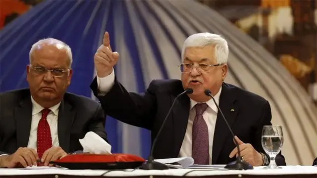 محمود عباس، رئیس تشکیلات خودگردان فلسطینی، در حالی که پشت میزی نشسته و مقام دیگری کنار او حضور دارد، با حالتی از دلخوری و عصبانیت دست خود را بالا برده است